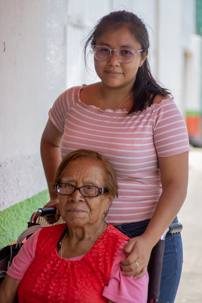 hero-img-01 A young woman supports an elderly lady in a wheelchair, showcasing care and compassion outdoors.