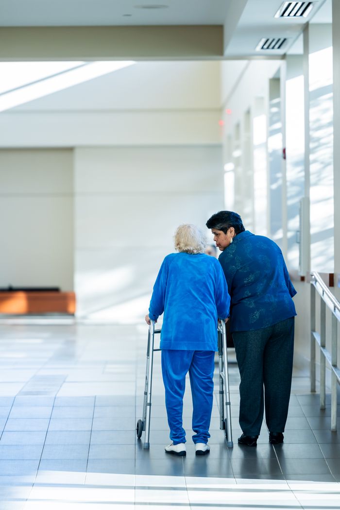 gallery-5 A nurse assists an elderly woman with a walker in a bright hospital corridor.