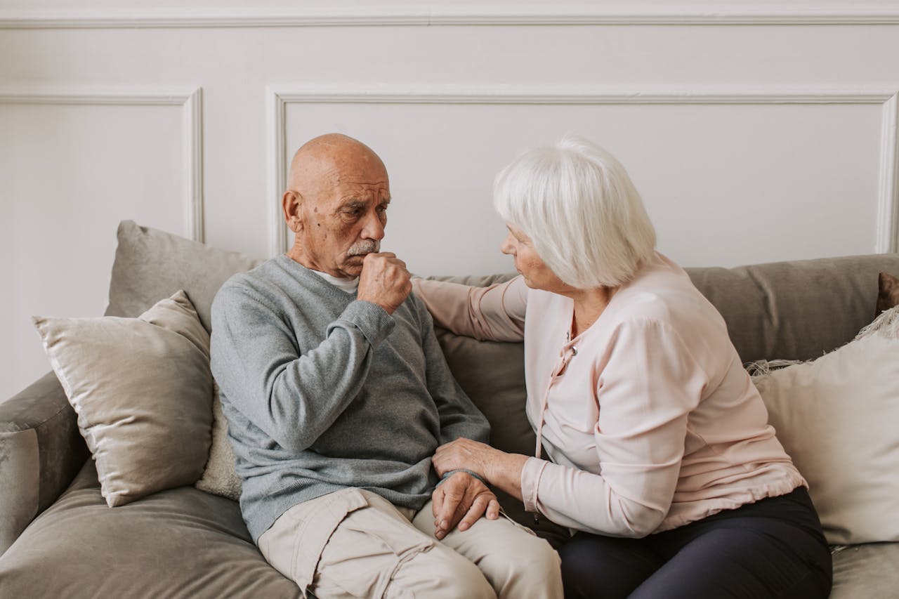 services-02 An elderly woman comforts a man coughing on a couch, showcasing care and affection.