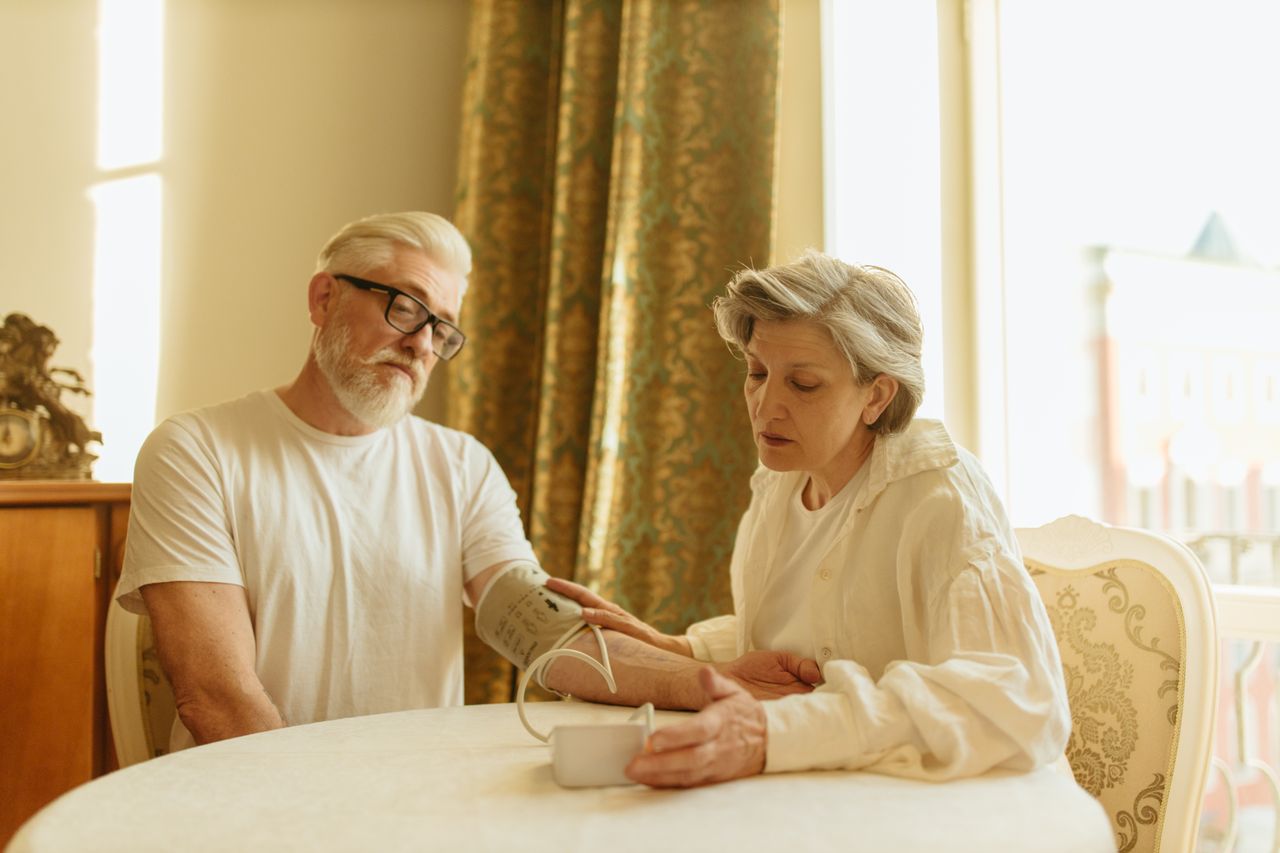 about-01 An elderly couple at home measuring blood pressure with a digital monitor, depicting care and health awareness.